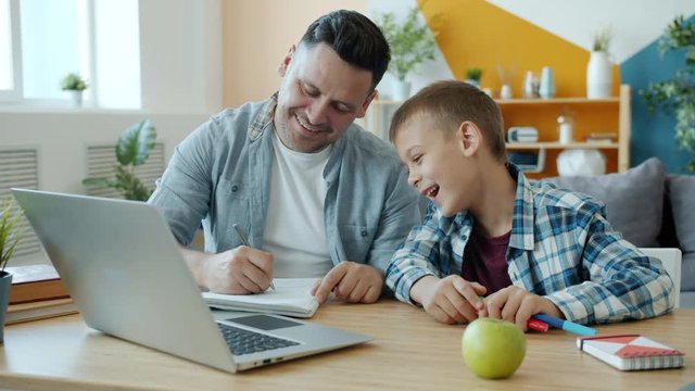 Young adult man is drawing in notebook having fun with happy child at home sitting at table with laptop together. Indoor activities and family concept.