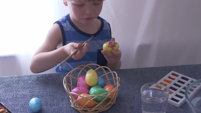 Little blond kid boy coloring eggs for Easter holiday in bath room, indoors. Child holding basket with painted eggs. Child having fun and celebrating feast with easter toy bynny