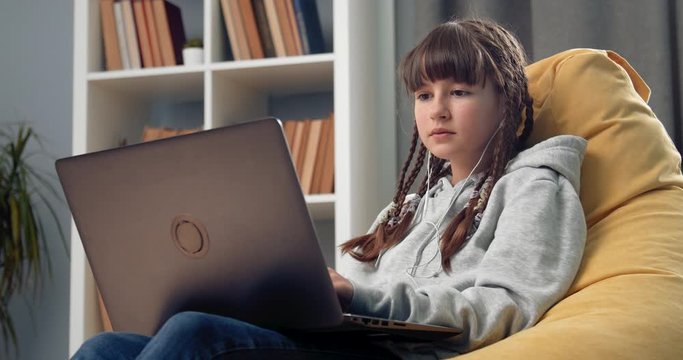 Young teenage girl with lots of braids sitting in modern yellow beanbag chair and using laptop. Pretty female in domestic outfit relaxing at home with digital gadget.