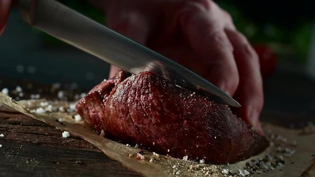 Man carving a small portion of roasted fillet beef steak seasoned with spices and condiments on a square of oven paper over a rustic wood counter Projekt