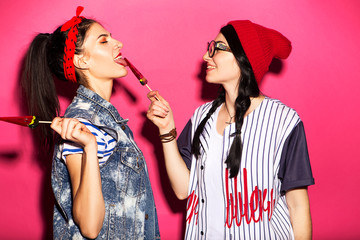 Two caucasian brunette hipster woman in casual stylish outfit having fun licking watermelon lollipops and playing with them. They standing on a bright pink background. Cheerful, happy emotions