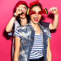 Two caucasian brunette hipster woman in casual stylish outfit having fun licking watermelon lollipops and playing with them. They standing on a bright pink background. Cheerful, happy emotions