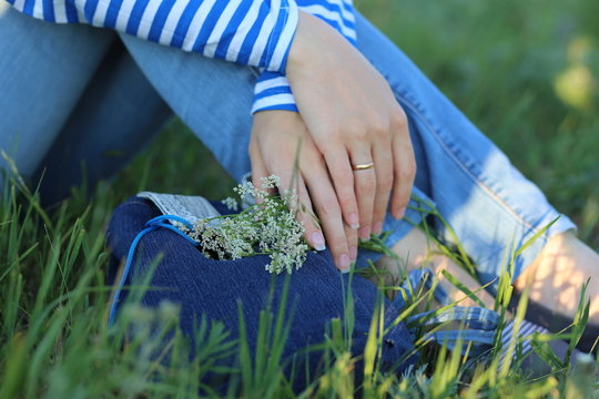 
the girl’s hands are holding white wildflowers on a denim bag, the girl is sitting on the green grass in blue jeans and a long sleeve t-shirt with blue and white stripes