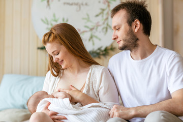 Young mother breast feeding her newborn child baby, father sits nearby and supports her