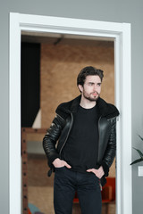 A young stylish man with a beard and dark hair in a black leather jacket with faux fur collar 