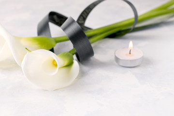 White Zantedesia flower tied with a black ribbon, next to a candle on a light background. Copy space. The concept of death, mourning
