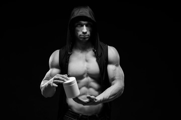 Naklejka na ściany i meble Muscular bodybuilder with jar of protein on a dark background. Sports nutrition. Bodybuilding nutrition supplements, sport, workout, healthy lifestyle concept. Black and white photography