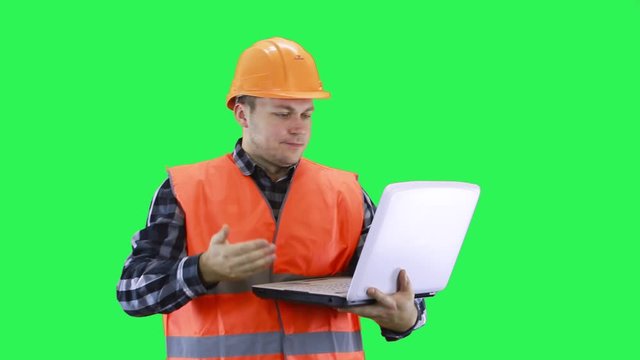 A man in a construction helmet and an orange vest communicates on a laptop making excuses in front of the interlocutor, green screen background