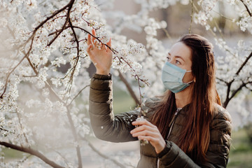 A girl with a medical mask on her face, allergic to flowers, stands in the garden of flowering cherry trees on a warm spring spring
