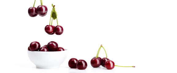 Ripe red cherries with green sticks and leaves fall into a white ceramic bowl.
