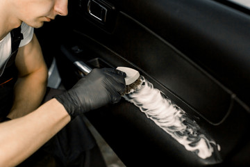 Cropped image of washing car interior by a soft brush with foam. Young male worker in black protective gloves cleaning car door inside with foam and brush