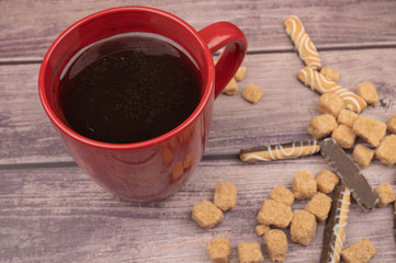 Red ceramic mug with tea, cookie sticks with chocolate and white icing, and pieces of brown cane sugar on a wooden background. Close up.