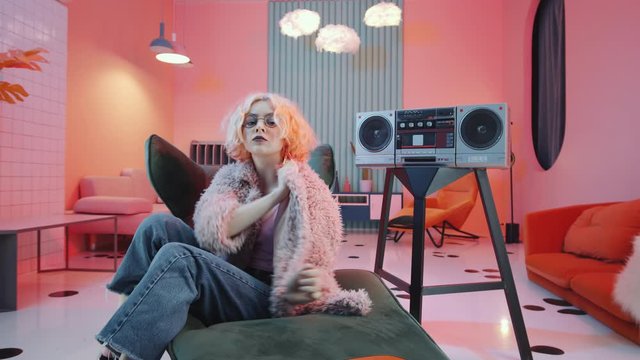Arc shot of sexy blonde woman in glamorous outfit performing vogue dance movements for camera on art deco armchair in vintage studio with pink lighting and retro cassette player
