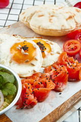 Unusual breakfast. Fried eggs, cherry tomatoes, guacamole hummus and salmon with pita on a board on a white background.