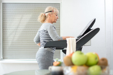 Active middle aged woman in sportswear using treadmill at home. Coronavirus Covid19 social distance. Home workout, active seniors, stay at home concept. - obrazy, fototapety, plakaty