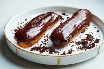 Two delicious eclairs with custard and glossy chocolate icing on a white ceramic plate on a marble background. Appetizing dessert and pastries. CLose up, selective focus