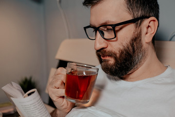 A lone bearded dark-haired man in glasses is drinking tea and reading a book while lying on his bed in the evening. Close-up on the right, cozy evening, bedroom, self-isolation and quarantine.