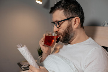 A lone bearded dark-haired man in glasses is drinking tea and reading a book while lying on his bed in the evening. Close-up on the right, cozy evening, bedroom, self-isolation and quarantine.