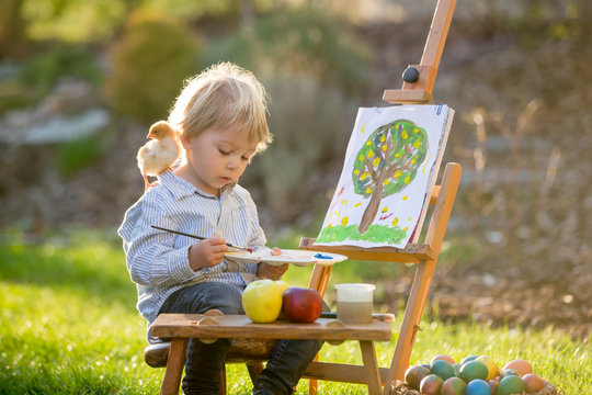 Sweet toddler child, painting eggs in garden with little chicks running around him