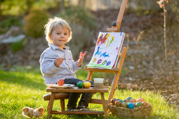 Sweet toddler child, painting eggs in garden with little chicks running around him