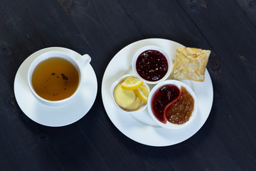 white cup of herbal tea with  plates of lemon, ginger, cranberry, raspberry, apple jam, baked stuffed ban on  black wooden background