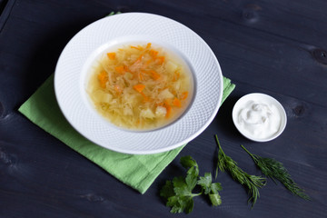 White plate with cabbage vegetarian soup on table cloth, sour cream, dill and parsley  on black wooden background