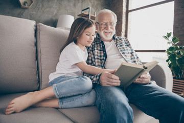 Photo of aged old grandpa little pretty granddaughter sit sofa hugging stay house quarantine safety read interesting book fairy tale modern interior living room indoors