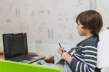 Distance learning for children during the coronavirus epidemic. The boy sits on the bed and receives a school assignment using the Internet and a laptop.