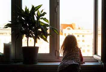 Girl sits on windowsill and looks out window. Stay at home. Child on home quarantine.  Kind sitz am...