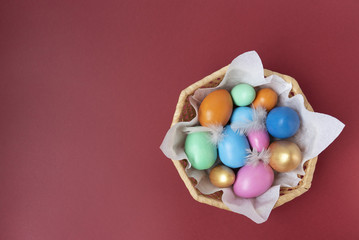 Fototapeta na wymiar Golden, pink, blue, green, orange eggs in the wicker basket on the maroon background. Copy space. Place for text and design. Happy Easter. Top view. Flat layout.