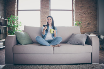 Full body photo of cheerful cute girl have covid-19 quarantine rest relax free time sit divan legs crossed watch tv switch channels enjoy hold cacao coffee cup in house indoors
