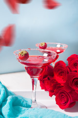 Red exotic alcoholic cocktail in clear glasses and red rose petals on the wooden white table for romantic dinner.