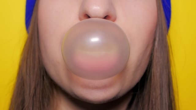 Portrait of a young sexy adult girl in a bright blue hat standing and isolated over a yellow background blowing chewing gum candy bubbles and chewing gum. A woman is looking at the camera. 4k. Slow
