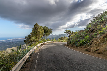 Panoramic view of asphalt road on the island coast surrounding hills around and see in a background Gran Canary island