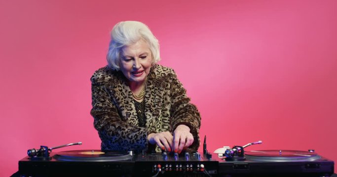 Dancing vintage retired Caucasian woman with grey hair and leopard coat and spinning record as DJ at retro party on pink wall background. Happy grandma having fun and playing music at disco of 80s.
