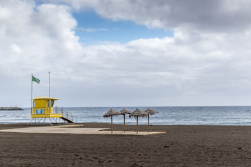 View of lifeguard tower on beach during windy sunny day with sea view and unique beach which has black sand on tower flutters green and yellow flag.