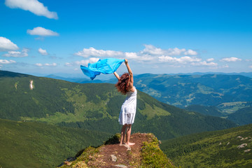 Young woman in white summer dress stands on a rock and looks into the valley, in her hands develops a lightweight blue scarf