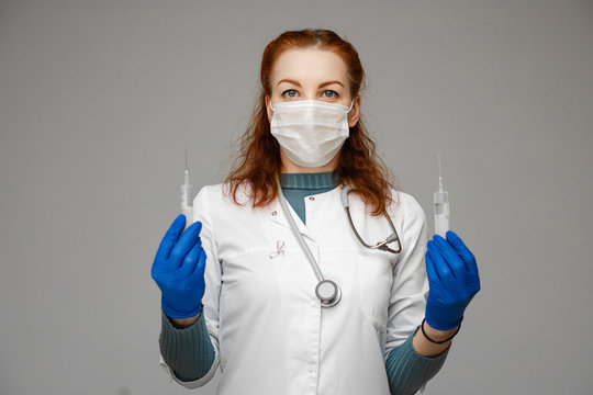 Stock photo portrait of a red-haired scientist in white coat, blue gloves and aseptic mask holding two syringes with treatment from coronavirus infection novel coronavirus disease 2019,COVID-19,nCoV