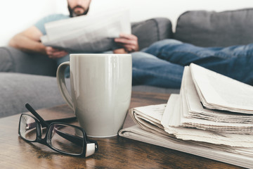 man relaxing on sofa reading a newspaper with stack of newspapers and coffee mug in foreground