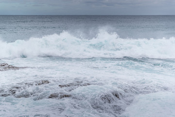 Waves crashing into a rocky shore full of large rocks and breaking water leaping to the sides with a pair of boats moored on the horizon at sea