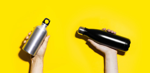 Close-up of male hands holding two steel and aluminum thermo bottles for water, black and silver of color. Isolated on yellow background.