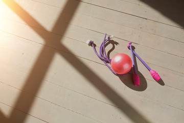 Pink gymnastic wands, pink ball and jump rope lying on the wooden floor in the rays of the rising...