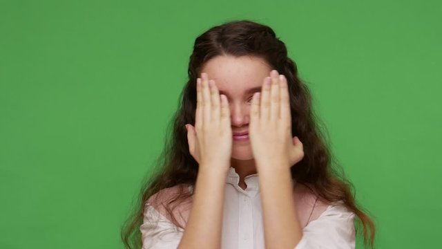 Closeup of carefree charming teenage brunette girl hiding face with hands, playing hide and seek game, peek a boo, changing grimaces and making faces. indoor studio shot isolated on green background