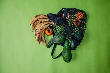 Mesh black reusable eco friendly string bag. Inside, delicious avocados, ripe bananas, persimmons, kiwi. The concept of recycling,environmental protection, naturalness. Greenish background. Copy space