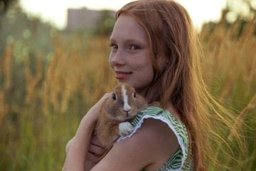 red-haired girl with freckles stands in a wheat field and holds a little rabbit