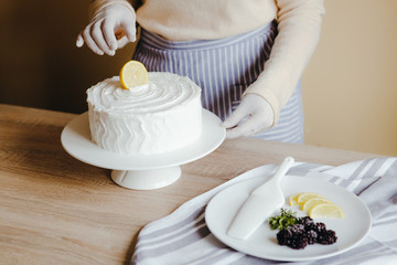 Housewife in the kitchen decorates a lemon cake with fruits, blackberry, lemon, mint during quarantine. Recipe of organic homemade lemon cake. Concept of cooking at home