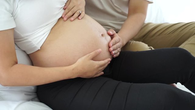 young asian  pregnant woman sitting on bed and husband touch the bally expecting baby with love during home quarantine from covid-19 virus pandemic