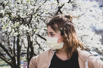 Attractive young girl wearing mask standing next to blooming cherry tree. Black t-shirtand beige top.April 2020