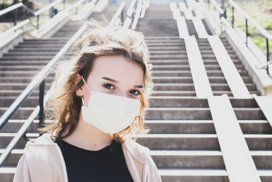 Outdoor Portrait Of Attractive Female With Brown Eyes And Brown Hair Wearing Hygienic Mask. Looking Seriously Into Camera.Black T-shirt, Beige Top.Urban Background With Backlight.Portait In Mask