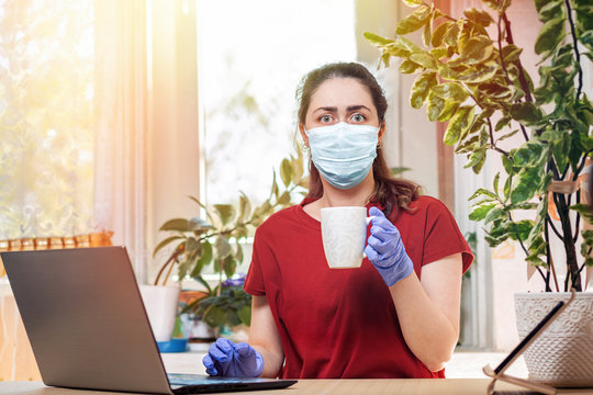 A Young Woman In Rubber Gloves And A Medical Mask With A Scared Look Holds A Mug In Her Hands. Home Decor, The Sun Outside The Window.The Concept Of Quarantine, Self-isolation And Remote Work